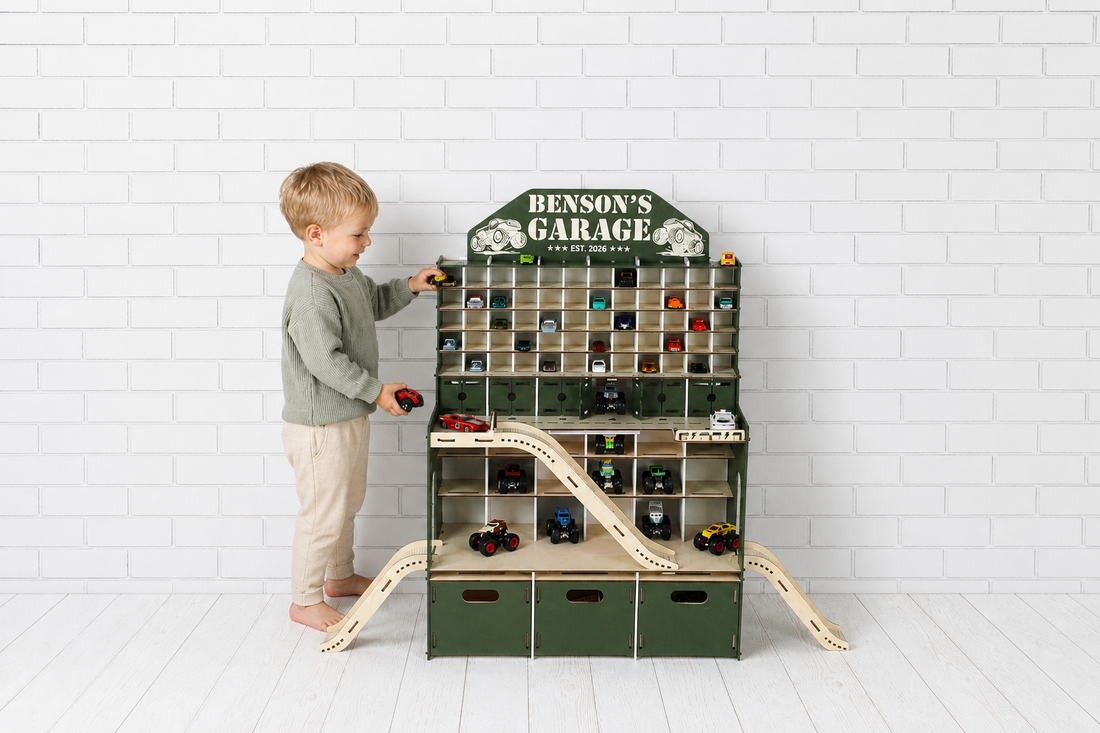 A young child plays with a personalized wooden toy car garage labeled “Benson’s Garage” in front of a white brick wall. The large green and natural wood garage features ramps, storage compartments, shelves, and small diecast toy cars arranged throughout.